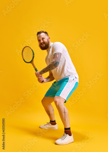 Full-length image of bearded man in sportswear posing with tennis racket against yellow studio background. Ready to play. Concept of sport, strength, fashion, emotions, lifestyle, ad © master1305