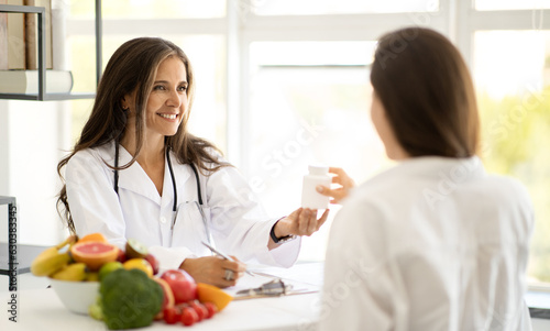 Cheerful mature caucasian lady doctor nutritionist in white coat advises young woman jar of vitamin pills at table