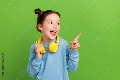 Photo of nice schoolgirl with buns dressed blue pullover in headphones directing look empty space isolated on green color background