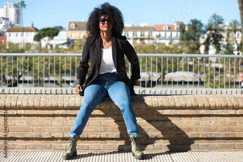 Young beautiful black woman with afro hair sitting on a bench wearing jeans and black jacket enjoying her holidays in spain. Travel and holiday concept.