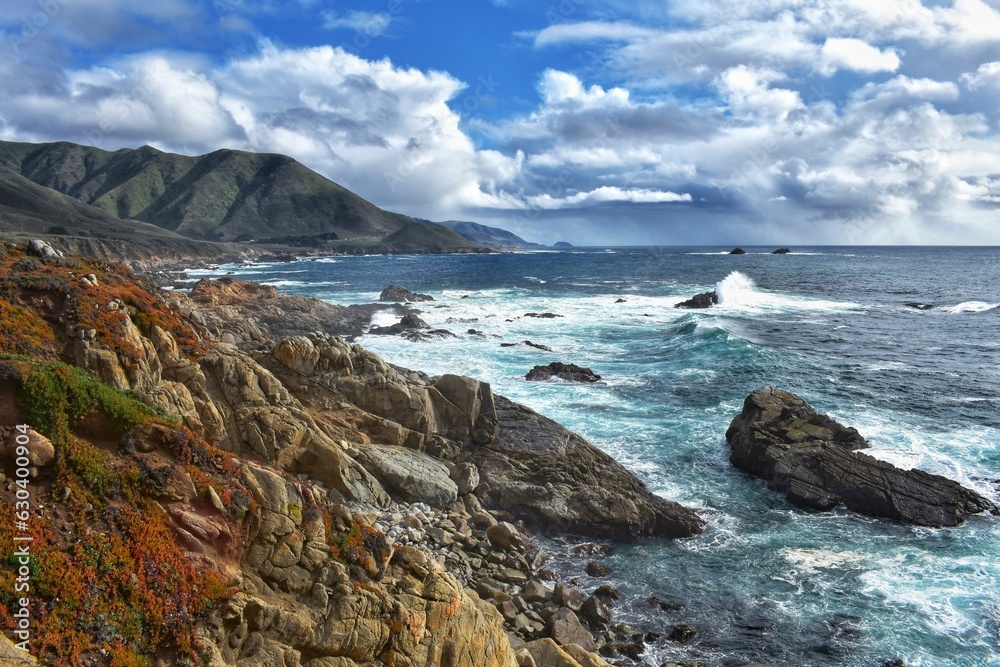 a rocky cliff face overlooking the ocean and mountain peaks on a bright day