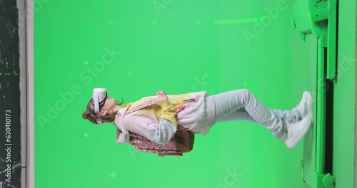 Vertical shot on green screen background. VR or AR online walk. Girl with a backpack in augmented reality googles walks through the virtual world and looks around. Walking on a treadmill side view.