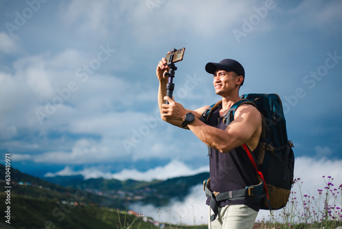 Tourist athletic man taking photo with smartphone and selfie stick on beautiful sky and fog background. Asian traveler blogger male with backpack enjoying shooting video on the top of mountains.