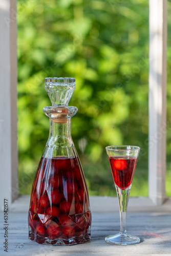 Homemade cherry brandy in wine glass and in a glass bottle on a wooden windowsill near summer garden, closeup