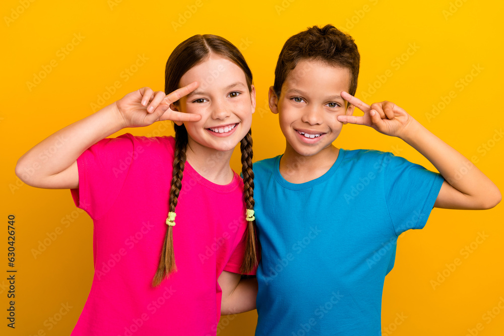 Photo of positive pretty preteen schoolkids dressed t-shirts showing v-signs cover eyes isolated yellow color background Photo of positive pretty preteen schoolkids dressed t-shirts showing v-signs cover eyes isolated yellow color background