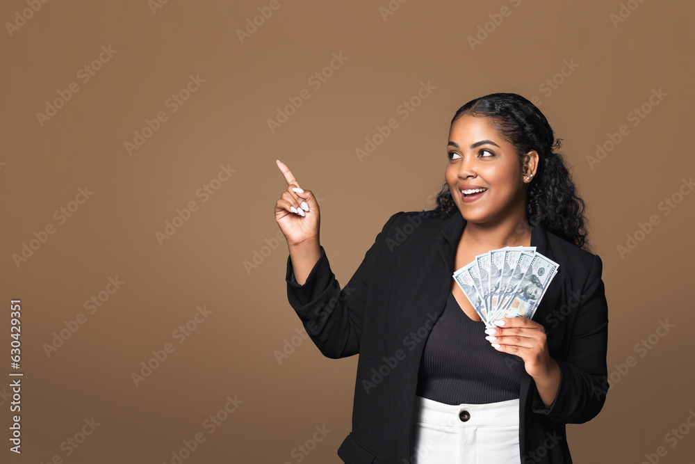 Happy latin plus size lady holding dollar cash fan in her hand and pointing aside at free space, brown studio background