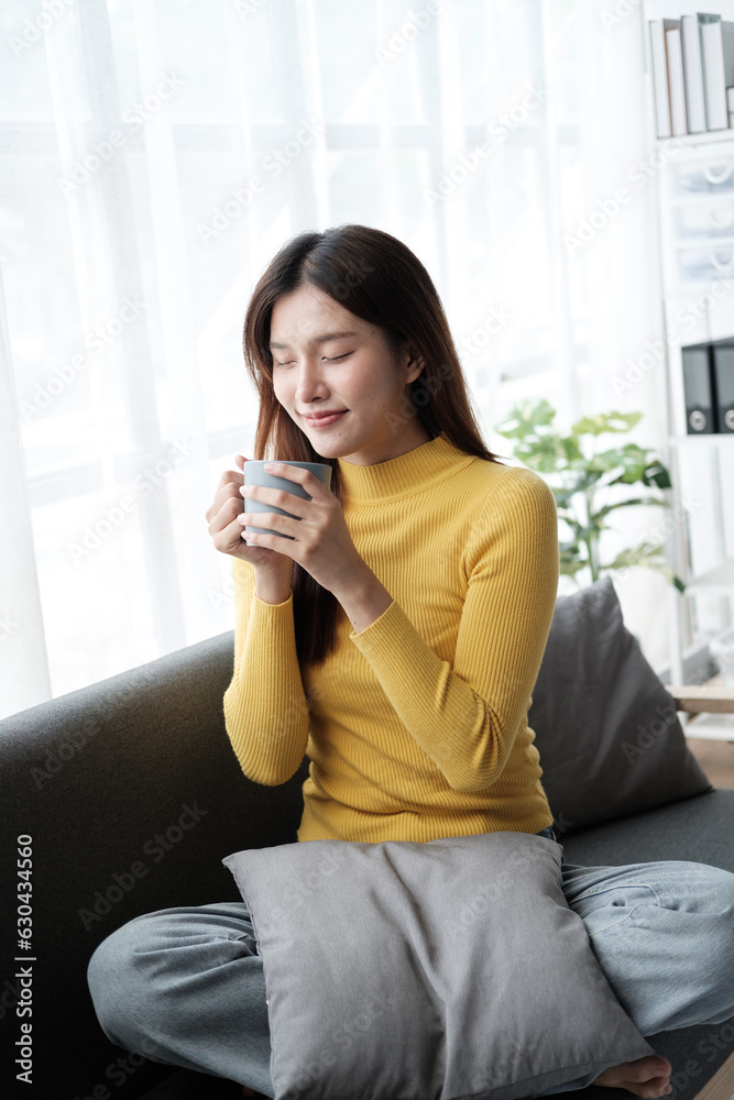 Young cheerful Asian woman sitting on the sofa with coffee cup in her hand, relaxing in her free tim at home. Young cheerful Asian woman sitting on the sofa with coffee cup in her hand, relaxing in her free tim at home.