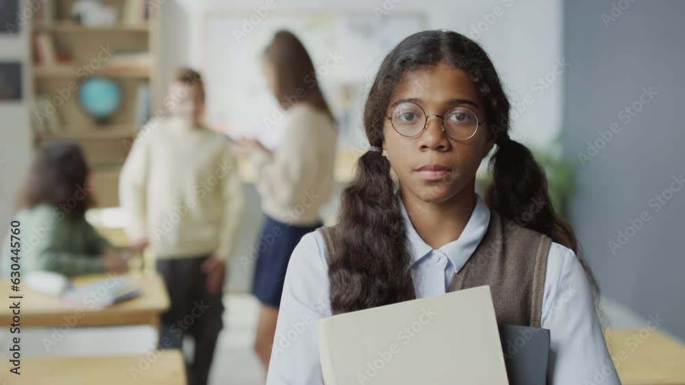 Selective focus medium close-up slowmo portrait of teenage Black girl wearing eyeglasses holding books looking at camera Selective focus medium close-up slowmo portrait of teenage Black girl wearing eyeglasses holding books looking at camera