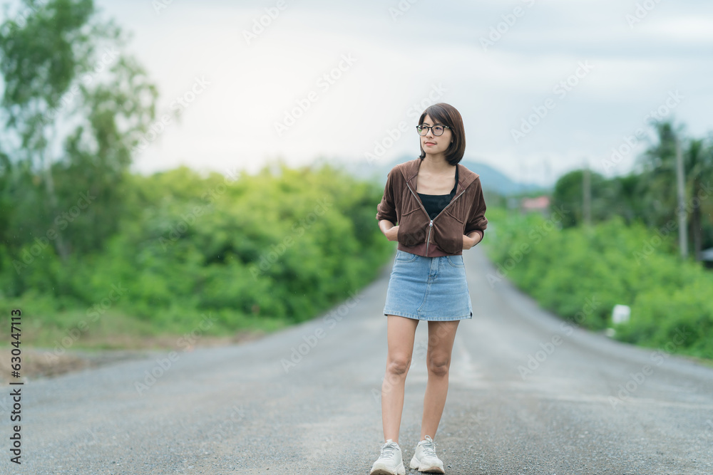 Asian woman wearing glasses standing on a tree-lined road behind there is a mountain