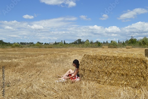 A girl with a jug in her hands sat down by a bale of hay in a mowed field on a sunny day.