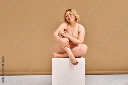 Cute adult blonde plus size woman in beige lingerie sits on a cube in a photo studio on a beige background and poses. Self-acceptance and self-love. photo