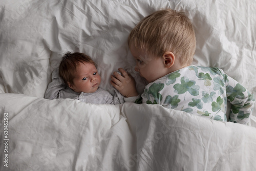proud toddler big brother boy holds hands with newborn baby sibling sister girl on white comforter blanket happy family in a bed  photo