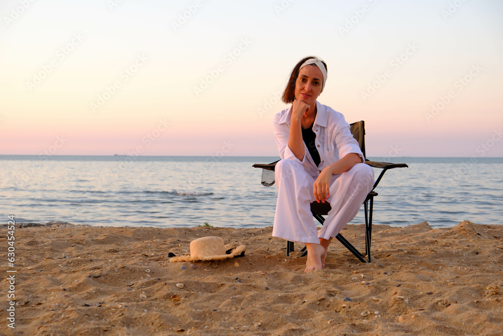Beautiful cute woman in white suit and hat is sitting and relaxing on sea beach coastline.