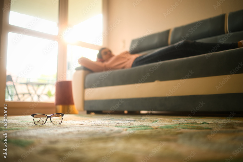 Close-up of glasses on the floor of the living room with young woman in the background laying on the sofa wearing a headset and listening to music relaxing and smiling in in warm sun light.