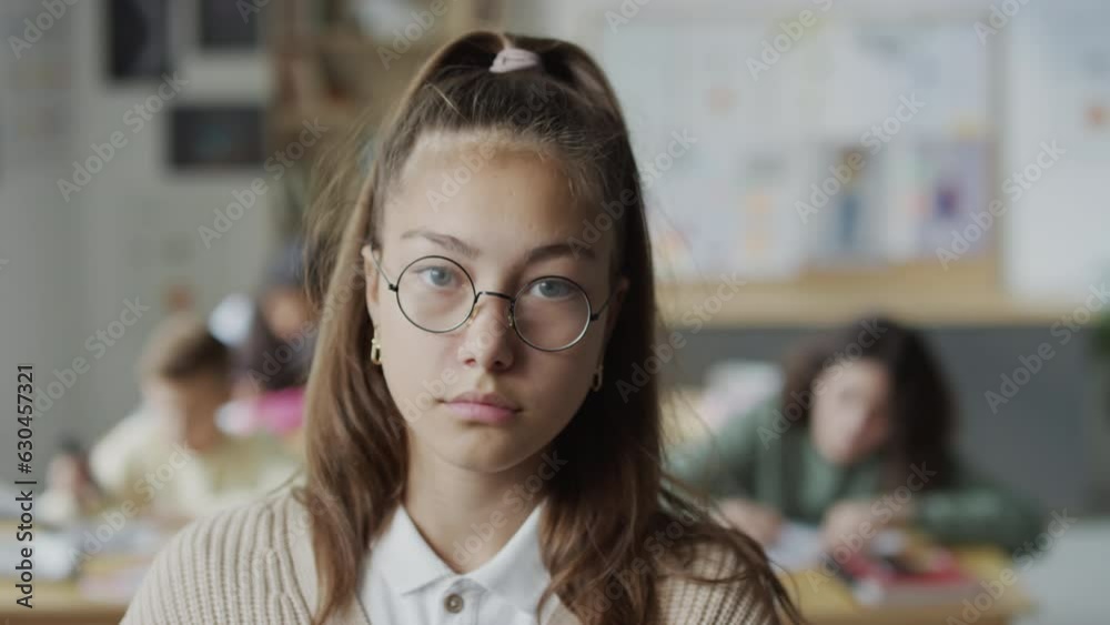 Selective focus close-up portrait of sad teen schoolgirl wearing eyeglasses standing in classroom looking at camera Selective focus close-up portrait of sad teen schoolgirl wearing eyeglasses standing in classroom looking at camera