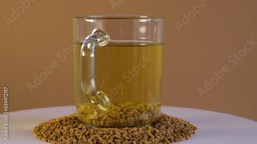 Cup of Egyptian fenugreek yellow tea, Helba or Methi Dana drink and fenugreek seeds on turntable, side view. 4K Video, Rotating. Brewed seeds used in alternative medicine. photo