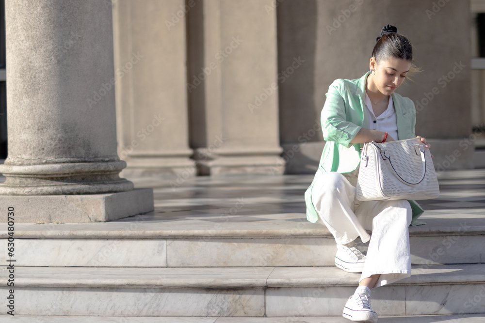 Young woman looking at her portfolio sitting on steps of public place. Dressed fashion style. Copyspace
