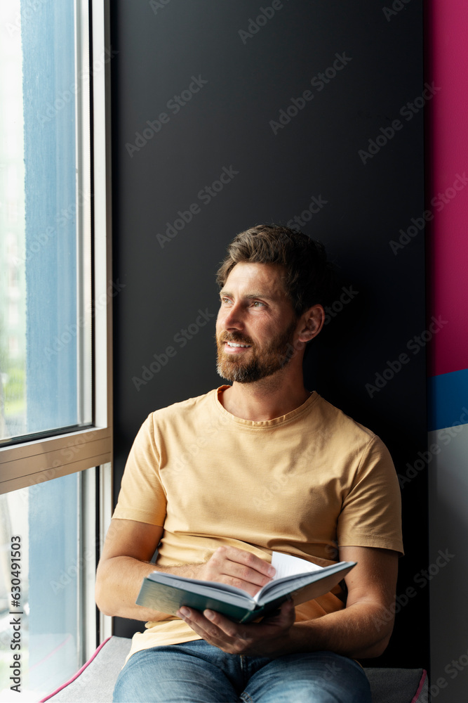 Portrait of handsome smiling man reading book sitting at home near window