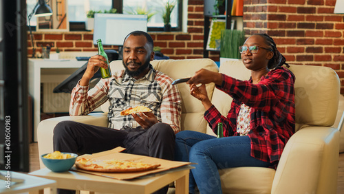 African american partners eating pizza slices at home, feeling happy watching film together. Relaxed couple having fun eating takeaway delivery food in living room, leisure activity.