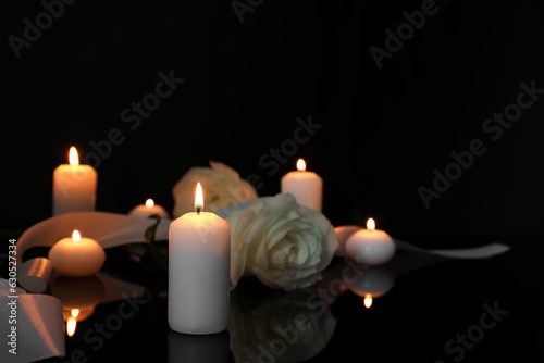 White roses and burning candles on black mirror surface in darkness  space for text. Funeral symbols
