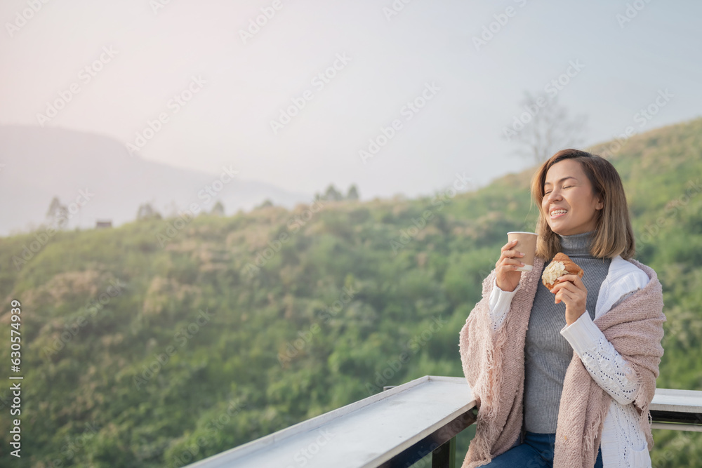 Fototapeta premium Relaxed woman breathing fresh air holding coffee cup and croissant for breakfast in autumn in a forest sitting on balcony and looking at mountains and green nature.Trekking concept..
