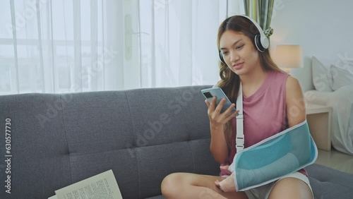 Young asian woman wearing soft splint due to sore arm using phone and listening music on sofa at home. photo