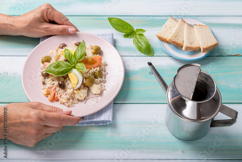 Female hands serve a plate of cold rice, topped with boiled eggs, olives, tomatoes, cheese and basil leaves