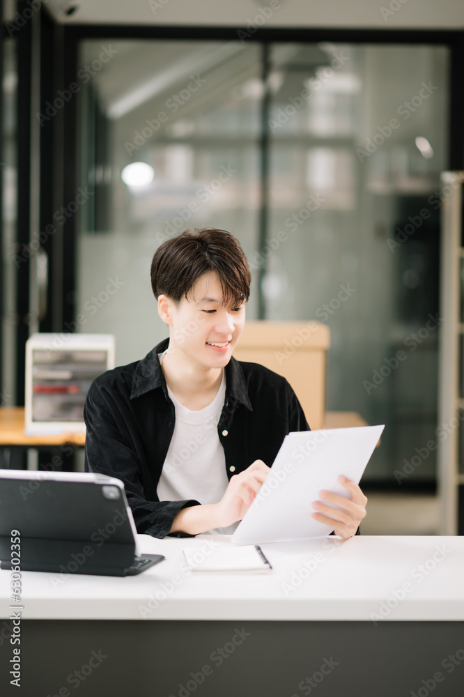 Young attractive Asian man smiling thinking planning writing in notebook, tablet and laptop working from home, looking at camera at office . Young attractive Asian man smiling thinking planning writing in notebook, tablet and laptop working from home, looking at camera at office .