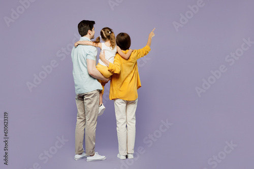 Full body back rear view young parents mom dad child kid girl 6 years old wearing blue yellow casual clothes hold daughter point finger aside isolated on plain purple background. Family day concept.