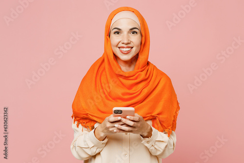 Photography Young arabian asian muslim woman wear orange abaya hijab hold in hand use mobile cell phone isolated on plain light pink background studio portrait