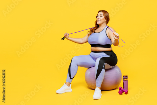 Full body young chubby plus size big fat fit woman wear blue top warm up training sit on fit ball hold in hand skipping rope isolated on plain yellow background studio home gym. Workout sport concept. © ViDi Studio