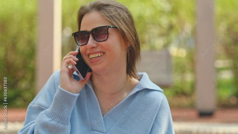 Smiling young woman wearing a blue sweater talking on phone sitting in a park Smiling young woman wearing a blue sweater talking on phone sitting in a park