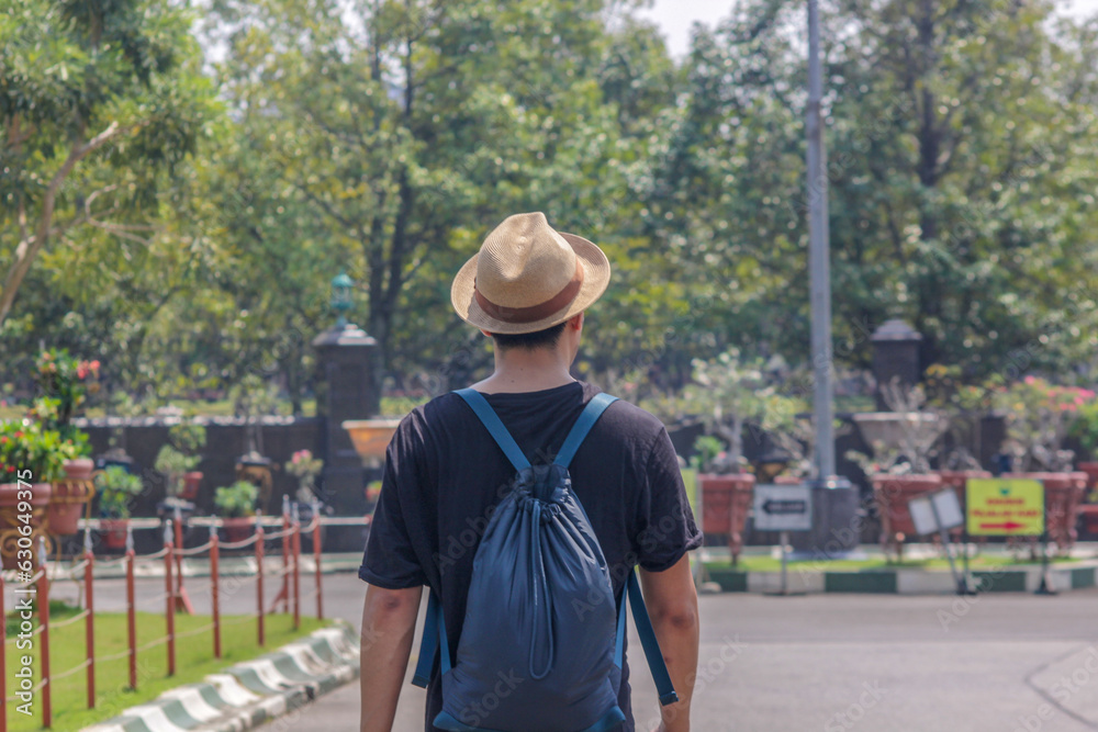 An Asian traveler wearing a hat and backpack is looking around from behind.