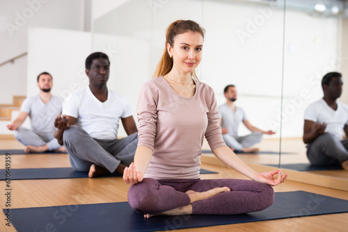 Young positive woman sitting in lotus pose on mat in fitness room, doing yoga meditation with group of adult people..
