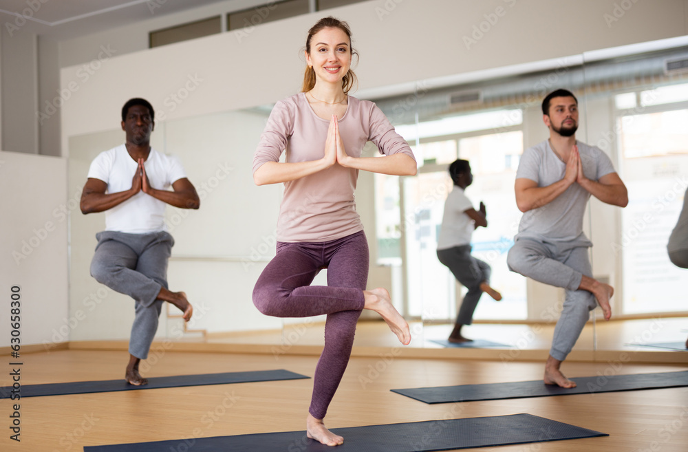 Full-length portrait of positive fit young woman standing on one leg in Eka Pada Utkatasana pose with clasped hands 