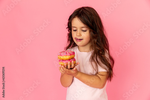 Portrait of a happy little smiling girl with curly hair and two appetizing donuts in her hands on a pink background, a place for text. Dieting concept and junk food. Side view