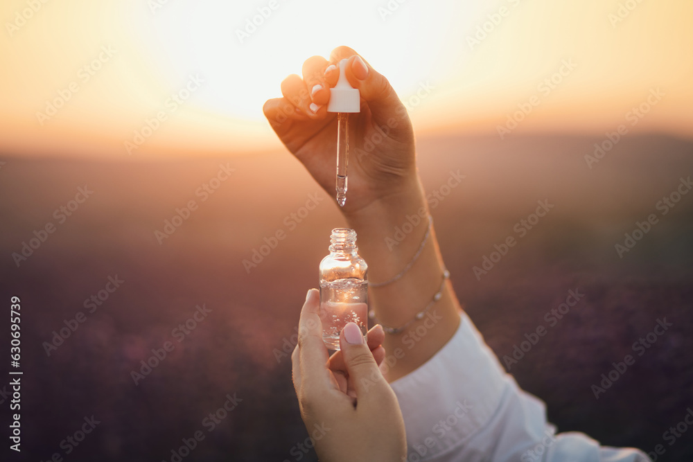 Close up female hands holding essential oil in a bottle with pipette in lavender field at sunset. - obrazy, fototapety, plakaty 