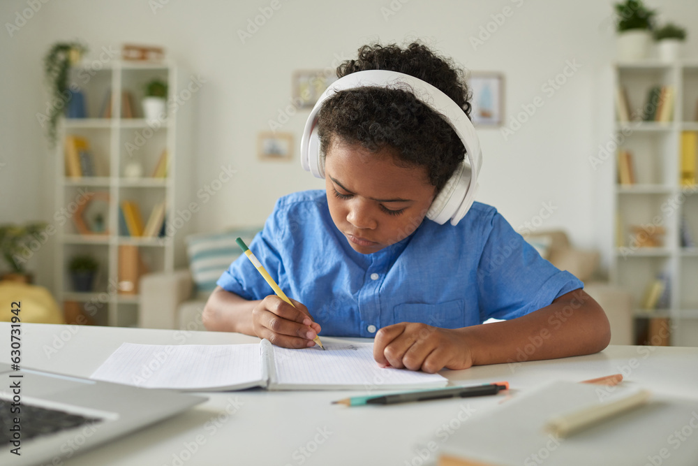 Serious African-American schoolboy in headphones sitting at desk and making notes while listening to tutor online Serious African-American schoolboy in headphones sitting at desk and making notes while listening to tutor online