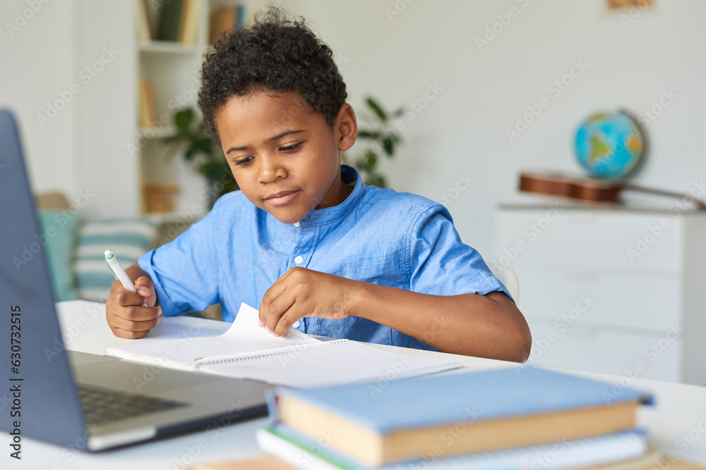 Curious black schoolboy with curly hair sitting at table and checking notes in workbook while doing homework Curious black schoolboy with curly hair sitting at table and checking notes in workbook while doing homework