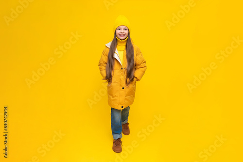 A little girl in full height in an autumn coat, down jackets and hats, rejoices in autumn. Autumn clothes for children. Yellow isolated background.
