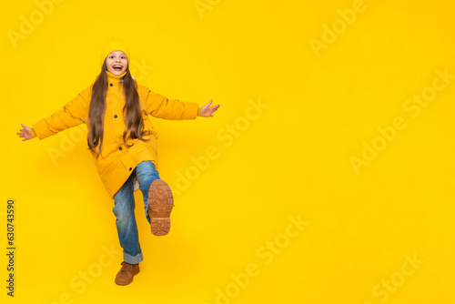 A little girl in full height in an autumn coat  down jackets and hats  rejoices in autumn. The child joyfully raises his leg with his arms outstretched to the sides. Yellow isolated background.