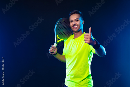 Padel tennis player celebrates victory in the game. Man athlete with paddle tenis racket on blue background. Sport concept. Download a high quality photo for sports website. Thumb up.