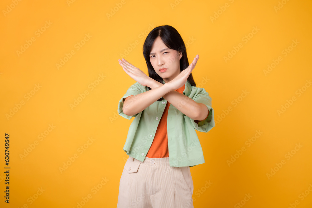 denial with a young Asian woman in her 30s, wearing an orange shirt and green jumper. Her cross hand gesture, isolated on a vibrant yellow background, conveys the concept of refusal and negation.