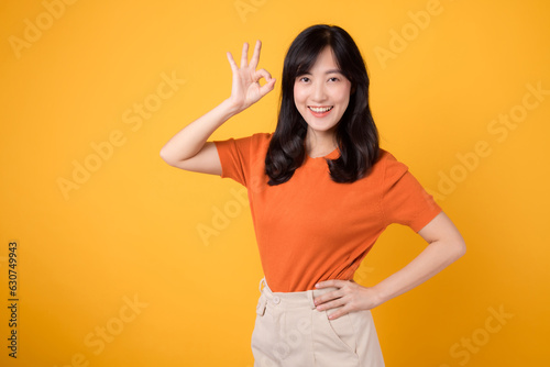 Confident young Asian woman in her 30s wears orange shirt, displaying okay sign on vibrant yellow background. Positive hand gesture concept. photo