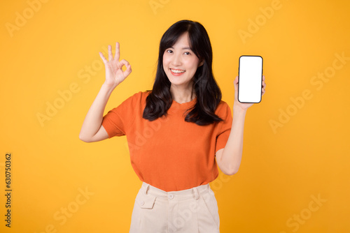 Energetic Asian woman in her 30s, wearing an orange shirt, shows okay gesture and smartphone blank display screen on yellow background. new application technology concept.