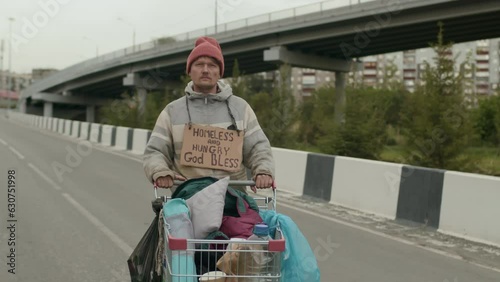 Portrait of homeless man with sign about need on his chest standing on empty highway and looking at cameraPortrait of homeless man with sign about need on his chest standing on empty highway and looki photo