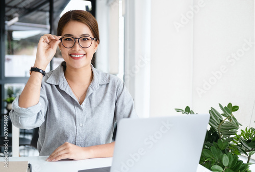 Joyful happy asian business woman in glasses sitting at workplace with laptop in home office, looking at camera, smiling. Millennial worker, employee, entrepreneur head shot portrait © Fahng