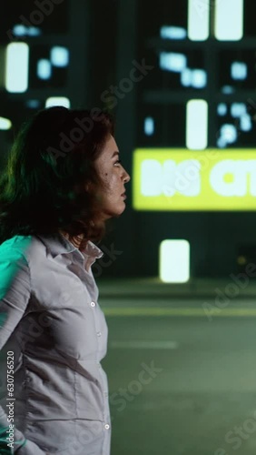 Vertical video Casual woman admires skyscrapes at night, walking around urban buildings and city lights on sidewalk. Entrepreneur strolling near downtown district with modern alleys and lamps, night photo
