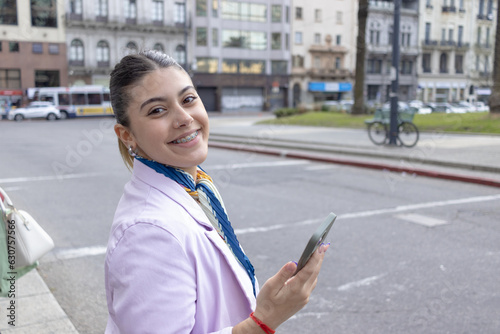 Young woman waits to cross the street with her smartphone in hand smiling at the camera. © martinkralicek