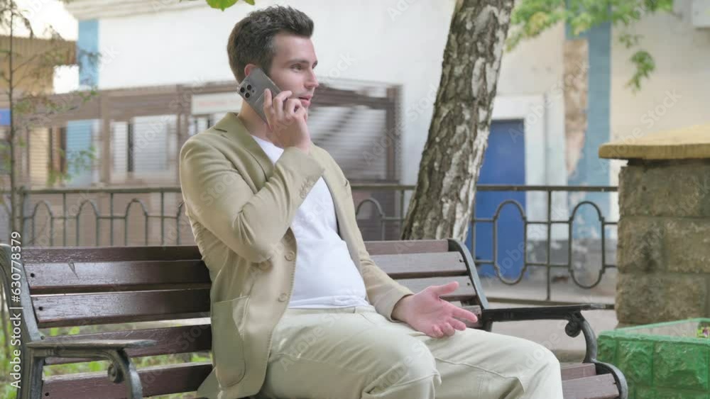 Young Man Talking on phone while Sitting Outdoor on a Bench Young Man Talking on phone while Sitting Outdoor on a Bench